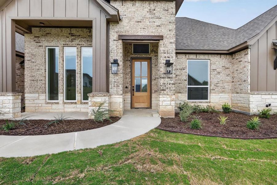 Brick and stone exterior featuring vertical panel siding accents, an inviting entryway with a wood-finish door, exterior wall lanterns, and a concrete pathway Brick and stone exterior featuring vertical panel siding accents, an inviting entryway with a wood-finish door, exterior wall lanterns, and a concrete pathway