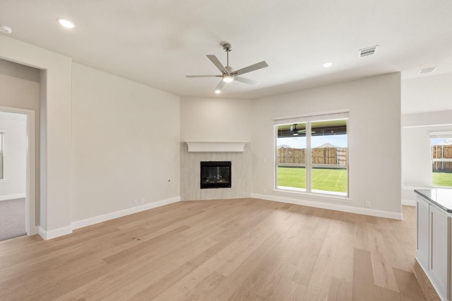 Open living room with light hardwood floors, tile fireplace, ceiling fan, and large backyard-facing window Open living room with light hardwood floors, tile fireplace, ceiling fan, and large backyard-facing window