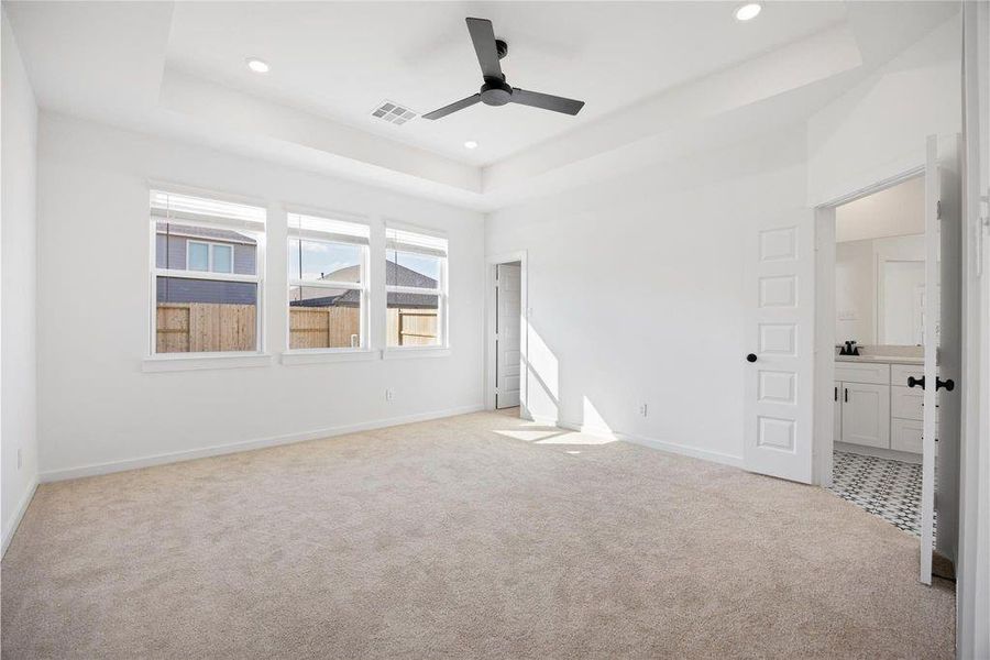 This spacious bedroom features plush carpet, large windows for natural light, a ceiling fan, and an attached bathroom with modern fixtures and patterned tile flooring. This spacious bedroom features plush carpet, large windows for natural light, a ceiling fan, and an attached bathroom with modern fixtures and patterned tile flooring.