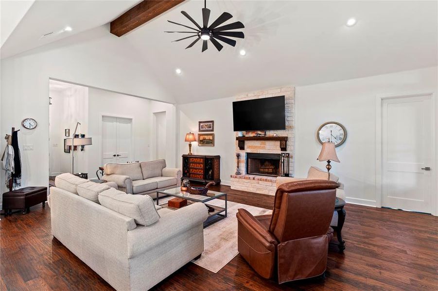 Living room with a stone fireplace, ceiling fan, beam ceiling, solid wood flooring, and high vaulted ceiling