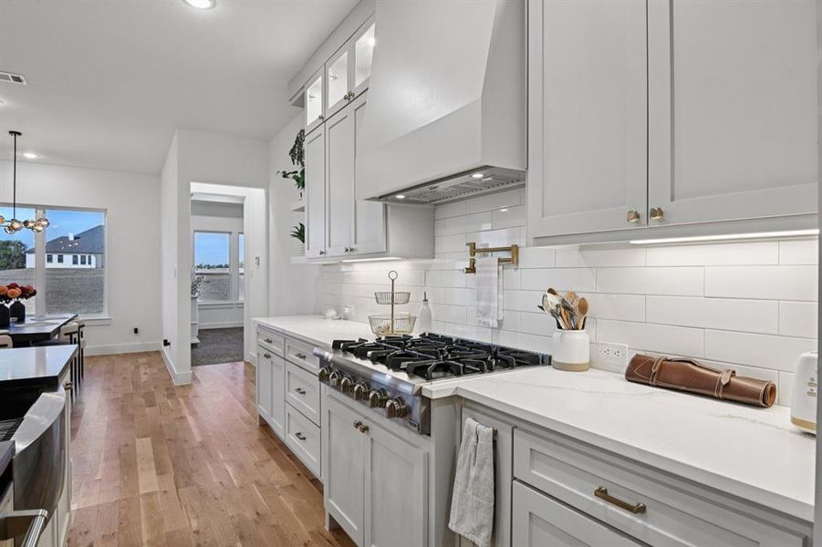 Kitchen featuring custom exhaust hood, stainless steel gas stovetop, light wood-style flooring, glass insert cabinets, and light stone counters Kitchen featuring custom exhaust hood, stainless steel gas stovetop, light wood-style flooring, glass insert cabinets, and light stone counters