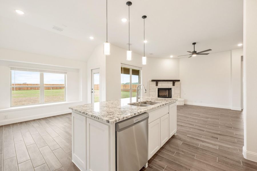 Kitchen island with granite countertop and stainless dishwasher, open to living room with brick fireplace and wood-look floors