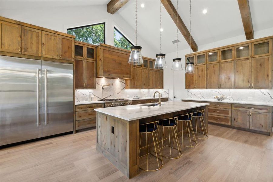 Kitchen with beamed ceiling, a center island with sink, appliances with stainless steel finishes, light stone counters, and light wood-type flooring