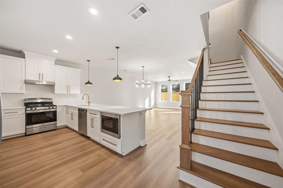 This modern home features an open-concept layout with a spacious kitchen, complete with white cabinets, stainless steel appliances, and an island. The adjacent living area is bright with natural light and has wood flooring throughout. The staircase adds an elegant touch, leading to the upper level.