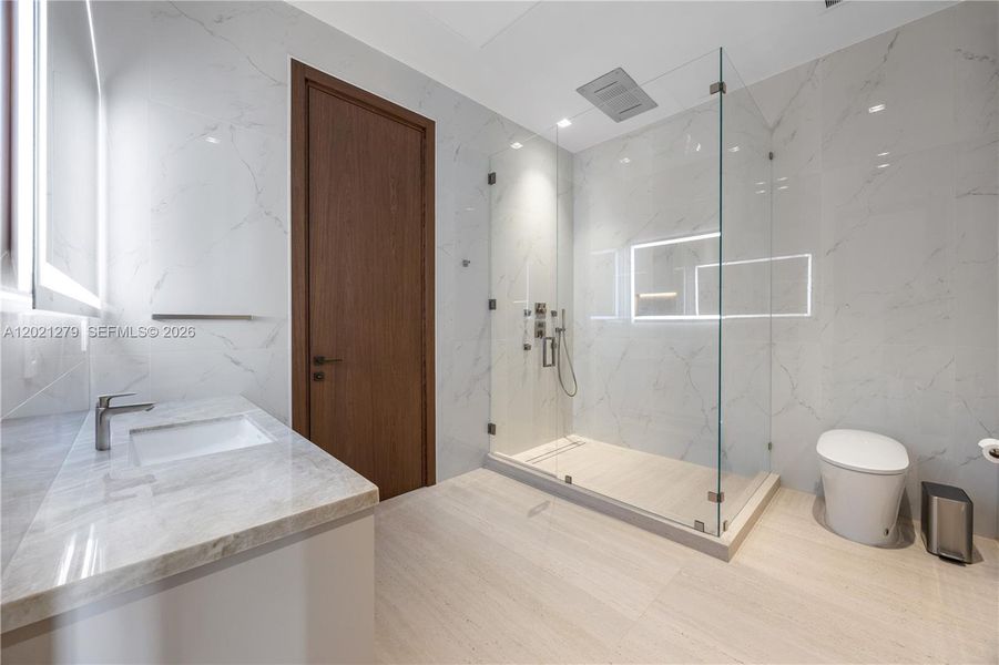 Modern guest bathroom with full-height white marble tile walls and floors, frameless glass walk-in shower, illuminated recessed niche, multi-function shower fixtures, undermount sink, stone vanity countertop, backlit mirror, and walnut door.