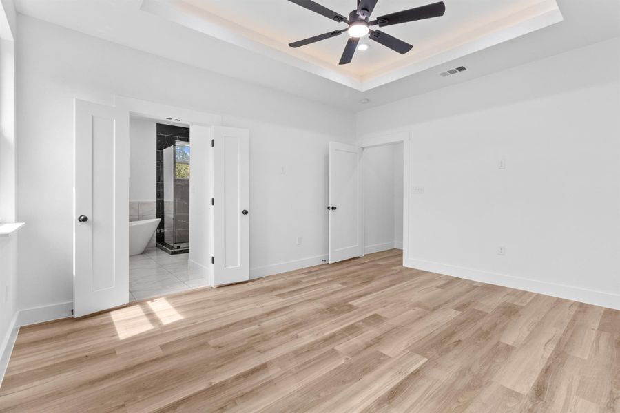 Unfurnished bedroom featuring a tray ceiling, light wood-type flooring, a ceiling fan, and ensuite bathroom