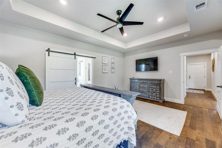Bedroom with a raised ceiling, a barn door, wood finished floors, recessed lighting, and ceiling fan