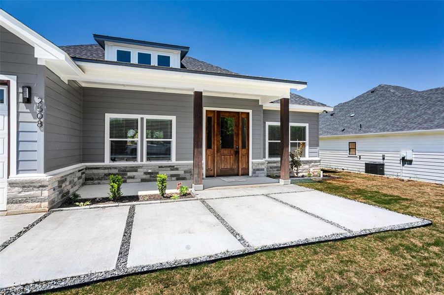 Entrance to property featuring roof with shingles, cooling unit, stone siding, and covered porch