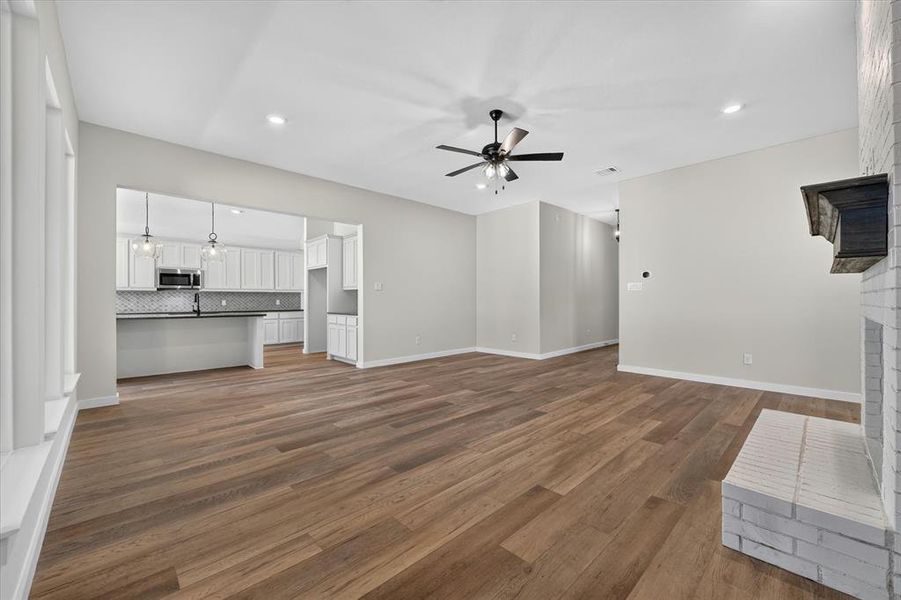 Spacious living area featuring wood-finish flooring, a white brick fireplace with a dark wood mantel, and an open concept layout to the kitchen Spacious living area featuring wood-finish flooring, a white brick fireplace with a dark wood mantel, and an open concept layout to the kitchen