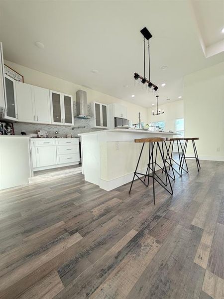 Side view of the kitchen with a stainless steel hood range, kitchen island with white quartz countertop, and luxury vinyl flooring.  White shaker self closing cabinets with hardware.  Custom backsplash with commercial / residential stainless faucet combo.