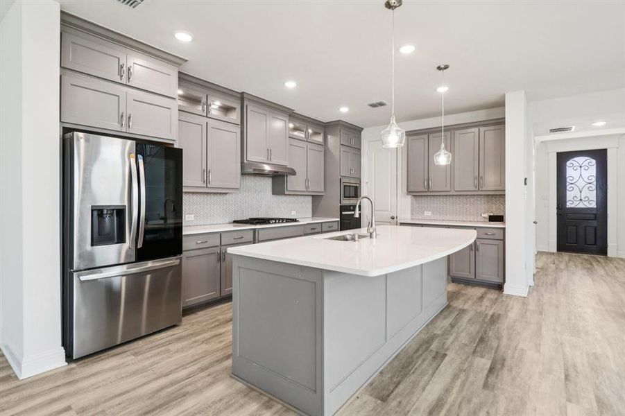 Kitchen featuring gray cabinetry, appliances with stainless steel finishes, a center island with sink, pendant lighting, and light wood-type flooring Kitchen featuring gray cabinetry, appliances with stainless steel finishes, a center island with sink, pendant lighting, and light wood-type flooring