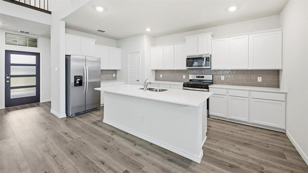 Modern kitchen featuring a central island with an integrated sink, white cabinetry, light countertops, stainless steel appliances, and a gray subway tile backsplash