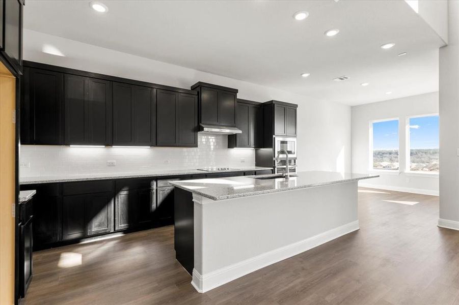 Kitchen with dark cabinetry, a center island with sink, light stone counters, dark wood-type flooring, and recessed lighting