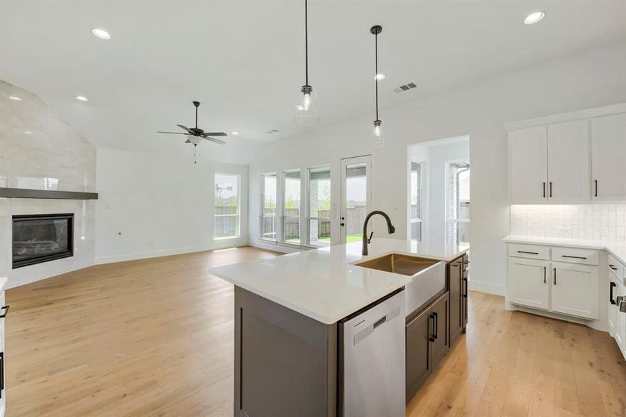 Open-concept kitchen and living area featuring a central island with a farmhouse sink, light wood-finish flooring, a contemporary fireplace with a stone surround, and multiple windows and glass doors providing natural light Open-concept kitchen and living area featuring a central island with a farmhouse sink, light wood-finish flooring, a contemporary fireplace with a stone surround, and multiple windows and glass doors providing natural light