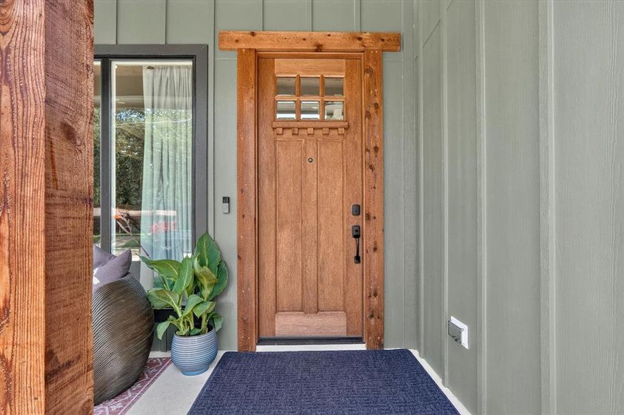 Craftsman-style wood entry door with transom window, framed by a timber post and light green board and batten siding