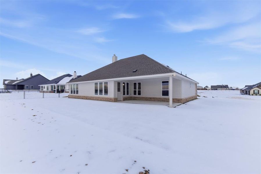 Snow covered house featuring a patio, a chimney, a shingled roof, and stone siding Snow covered house featuring a patio, a chimney, a shingled roof, and stone siding