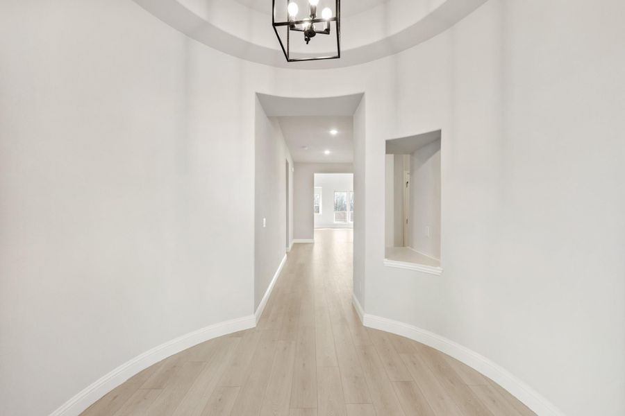 Curved entryway with round tray ceiling, black chandelier, light hardwood floors, and wall niche leading to hallway