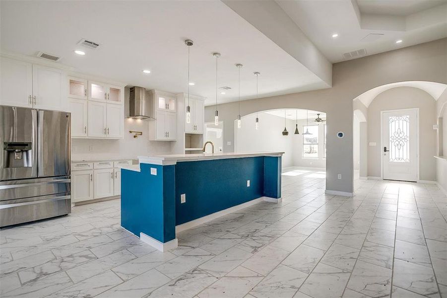 Kitchen with stainless steel fridge, wall chimney range hood, white cabinets, hanging light fixtures, and an island with sink