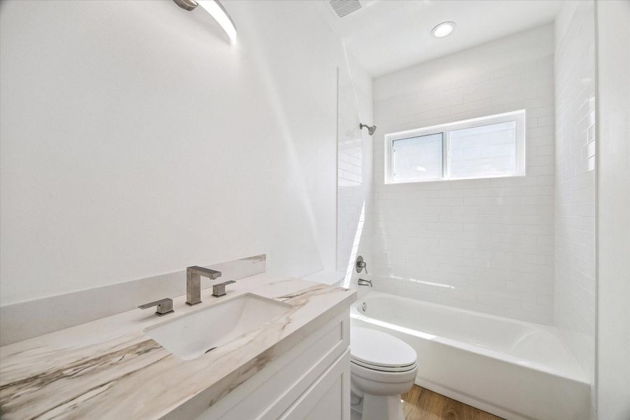 This bright full bathroom features a clean white vanity with a marble-look countertop, under-mount sink, and sleek modern faucet. The tub and shower combination is surrounded by glossy white subway-style tile extending toward the ceiling for a polished finish. A horizontal window introduces natural light, while recessed and vanity lighting keep the space bright and functional.