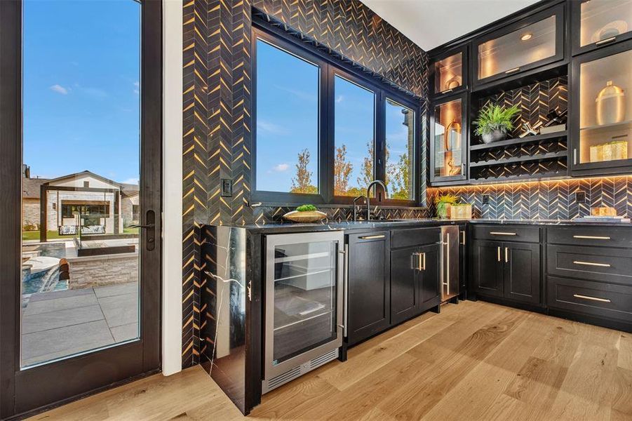 Indoor wet bar with dark cabinetry, wine cooler, light wood-style flooring, glass insert cabinets, and dark stone countertops Indoor wet bar with dark cabinetry, wine cooler, light wood-style flooring, glass insert cabinets, and dark stone countertops