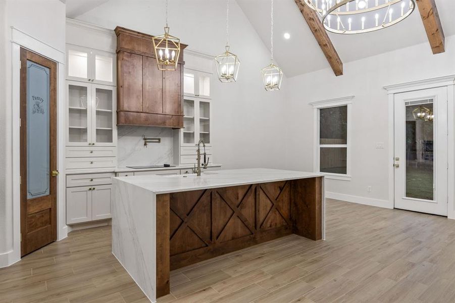 Kitchen featuring a two-tone island with a waterfall countertop, white cabinetry with glass-front uppers, a custom wood vent hood, and a pantry door with frosted glass Kitchen featuring a two-tone island with a waterfall countertop, white cabinetry with glass-front uppers, a custom wood vent hood, and a pantry door with frosted glass