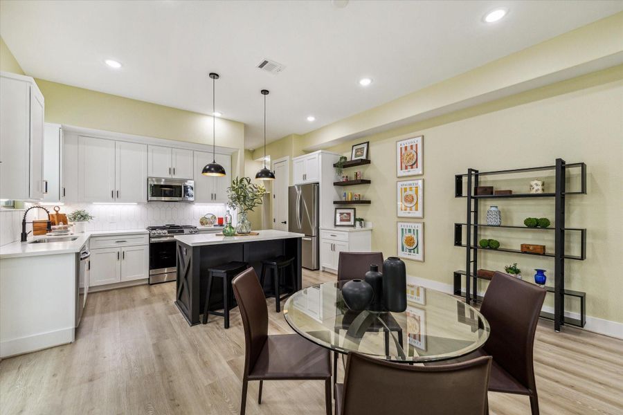 This modern kitchen features sleek white cabinetry, a central island with pendant lighting, and stainless steel appliances. The open layout includes a cozy dining area with a glass table and stylish shelving for decor. Perfect for entertaining.