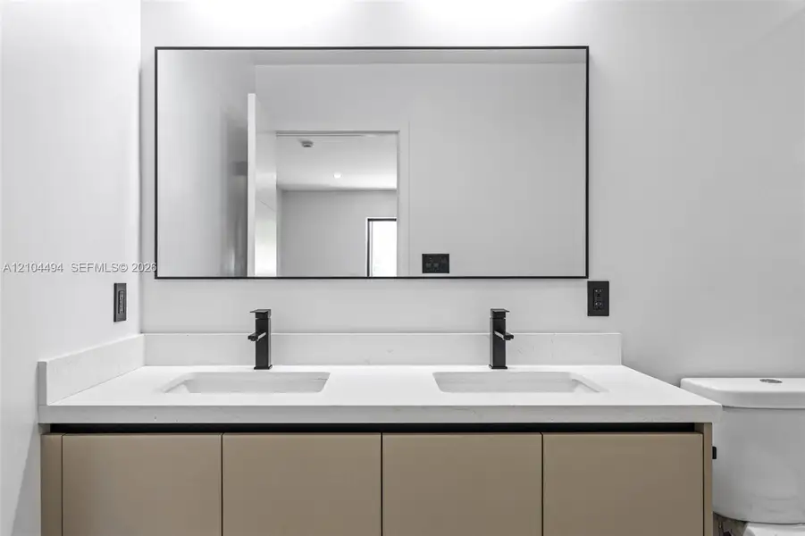 Double vanity featuring solid-surface countertop, dual rectangular undermount sinks, matte black widespread faucets, flat-panel cabinetry, and a large rectangular mirror with a thin black frame