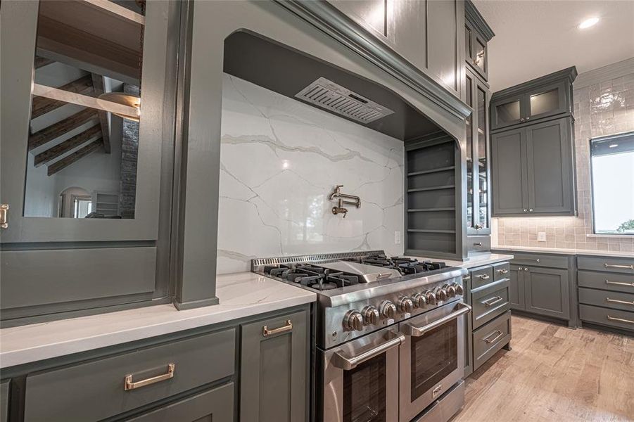 Kitchen with tasteful backsplash, range with two ovens, glass insert cabinets, light stone countertops, and gray cabinetry Kitchen with tasteful backsplash, range with two ovens, glass insert cabinets, light stone countertops, and gray cabinetry