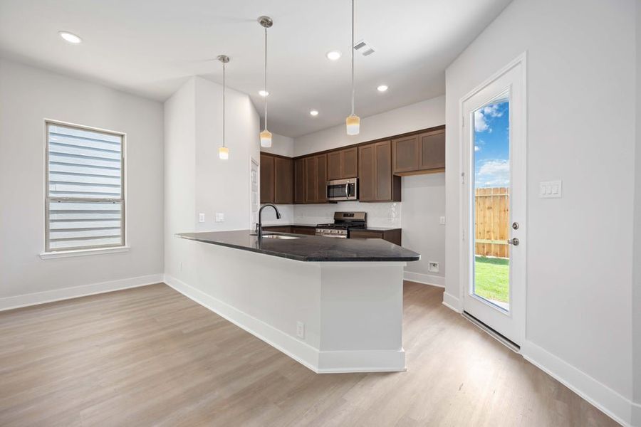 Open-concept kitchen with dark wood cabinets, gray countertops, pendant lights, and light wood flooring Open-concept kitchen with dark wood cabinets, gray countertops, pendant lights, and light wood flooring
