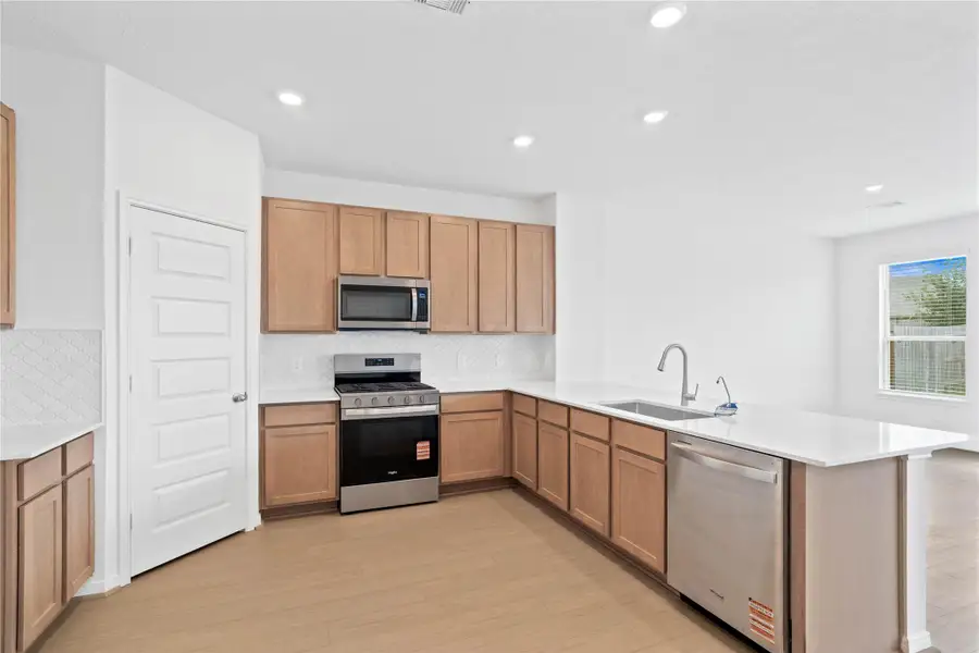 This spacious kitchen boasts high ceilings, Woodmont Dakota cabinets in a Barley stain, Silestone Lusso countertops, stainless steel Whirlpool appliances, a modern Daltile backsplash, recessed lighting, an extended breakfast bar countertop, and a pantry—all overlooking the expansive Great Room.