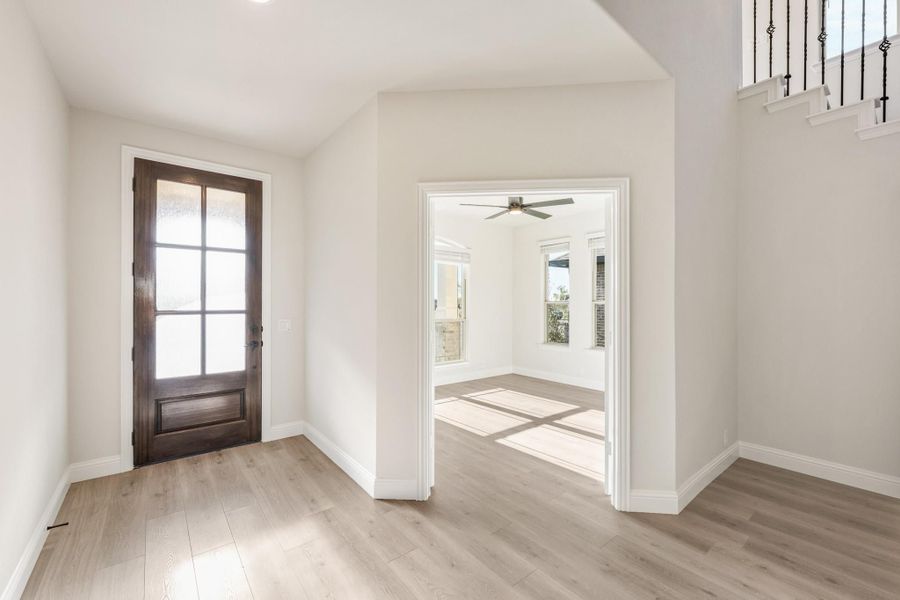 Entryway with dark wood glass-panel front door, light hardwood floors, and staircase railing leading to adjacent room with ceiling fan. Entryway with dark wood glass-panel front door, light hardwood floors, and staircase railing leading to adjacent room with ceiling fan.