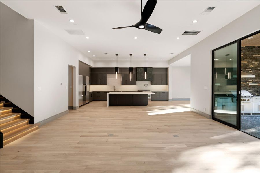 This modern open-concept living space features a sleek kitchen with dark cabinetry and a central island. The room is bright with natural light, enhanced by light wood flooring and recessed lighting. A stylish ceiling fan adds a contemporary touch, and sliding doors lead to an outdoor area, ideal for entertaining.
