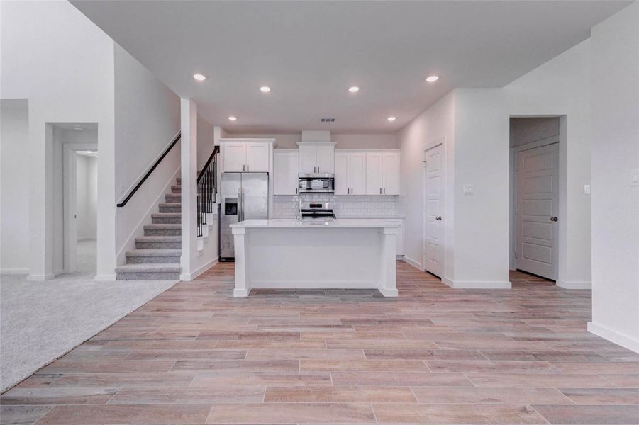Spacious kitchen and dining view with wood-look tile flooring, recessed lighting, white cabinetry, a central island, stainless appliances, and a clean open layout. The kitchen is positioned for easy everyday living, with the staircase nearby and the family room just beyond. Neutral finishes and black hardware provide a timeless, modern contrast throughout the space. Spacious kitchen and dining view with wood-look tile flooring, recessed lighting, white cabinetry, a central island, stainless appliances, and a clean open layout. The kitchen is positioned for easy everyday living, with the staircase nearby and the family room just beyond. Neutral finishes and black hardware provide a timeless, modern contrast throughout the space.