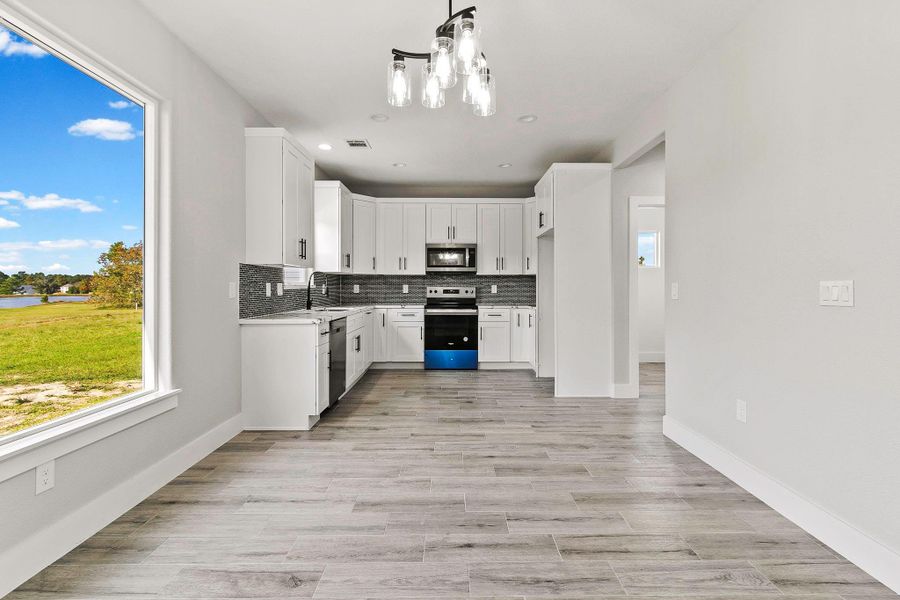 This photo showcases a bright, modern kitchen with white cabinetry, stainless steel appliances, and a stylish backsplash. The open space features wood-look flooring and a large window offering scenic views of greenery and a nearby lake. The contemporary lighting fixture adds a touch of elegance.