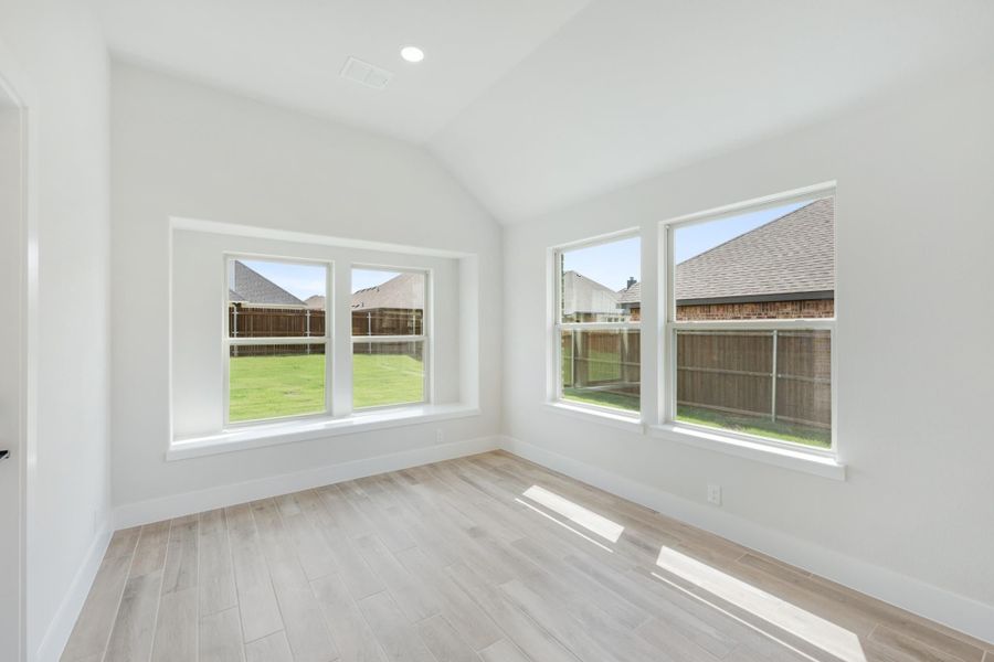 Empty room with vaulted ceiling, light wood floors, and large windows overlooking a fenced backyard