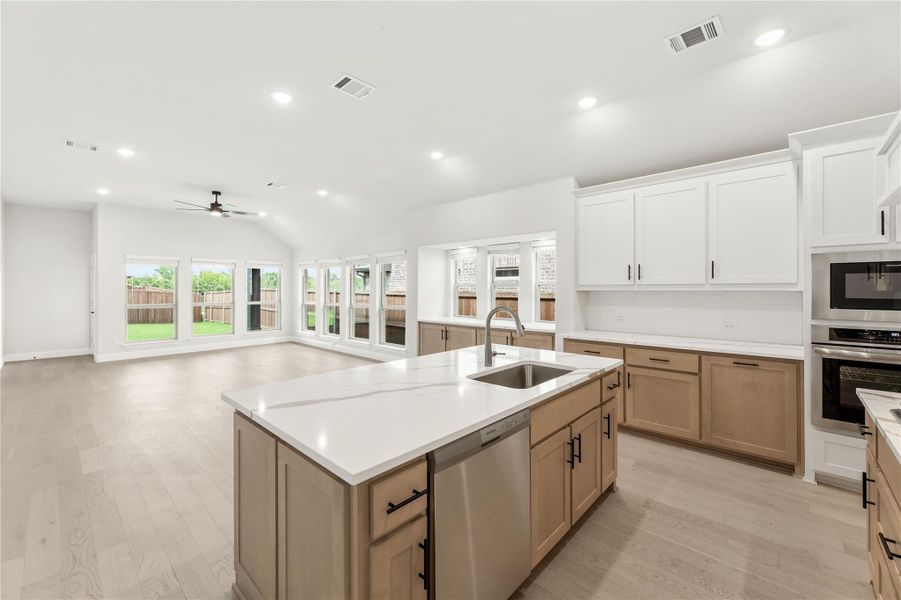 Open kitchen with large island, white quartz countertops, tan cabinets, stainless appliances, and light wood floors open to living area.