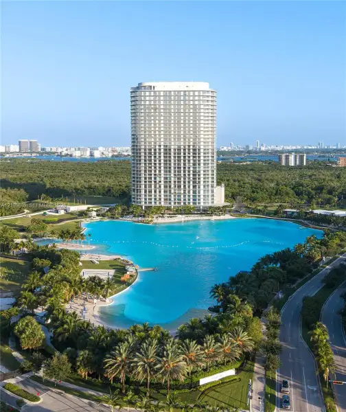 High-rise residential tower featuring floor-to-ceiling glass windows, a large man-made lagoon with sandy shorelines, and landscaped grounds with mature palm trees
