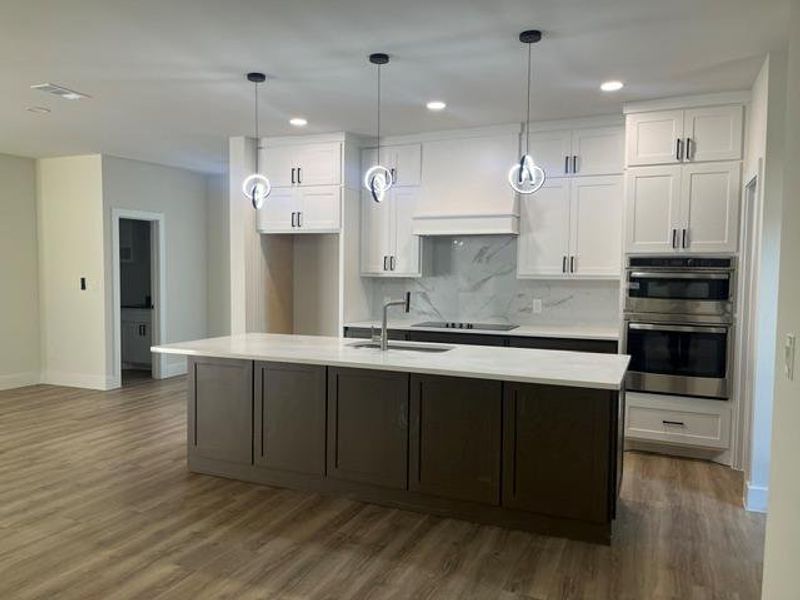 Spacious kitchen featuring an expansive center island with a light-toned countertop, white shaker cabinetry, stainless steel double wall ovens, a contemporary hood vent, and wood-finish flooring Spacious kitchen featuring an expansive center island with a light-toned countertop, white shaker cabinetry, stainless steel double wall ovens, a contemporary hood vent, and wood-finish flooring