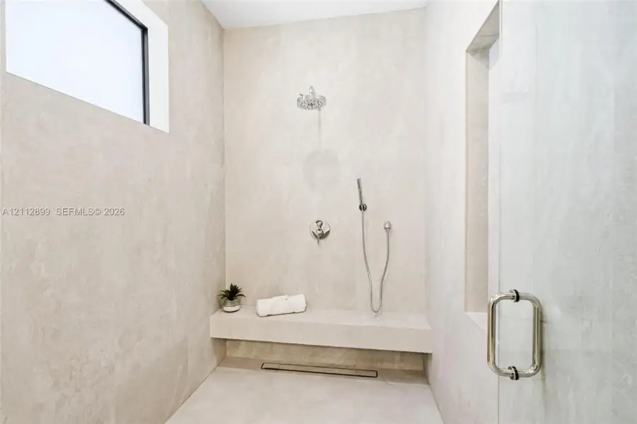 Walk-in shower featuring floor-to-ceiling stone-finish tile, a built-in stone bench, overhead rainfall showerhead, adjustable handheld shower wand, and glass enclosure door