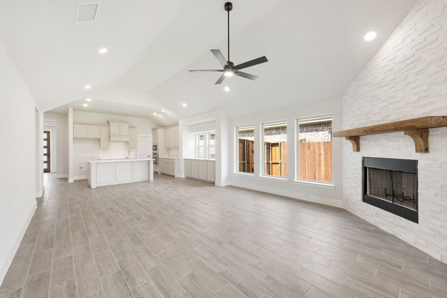 Open living area with vaulted ceiling, white brick fireplace, ceiling fan, and open kitchen with island.