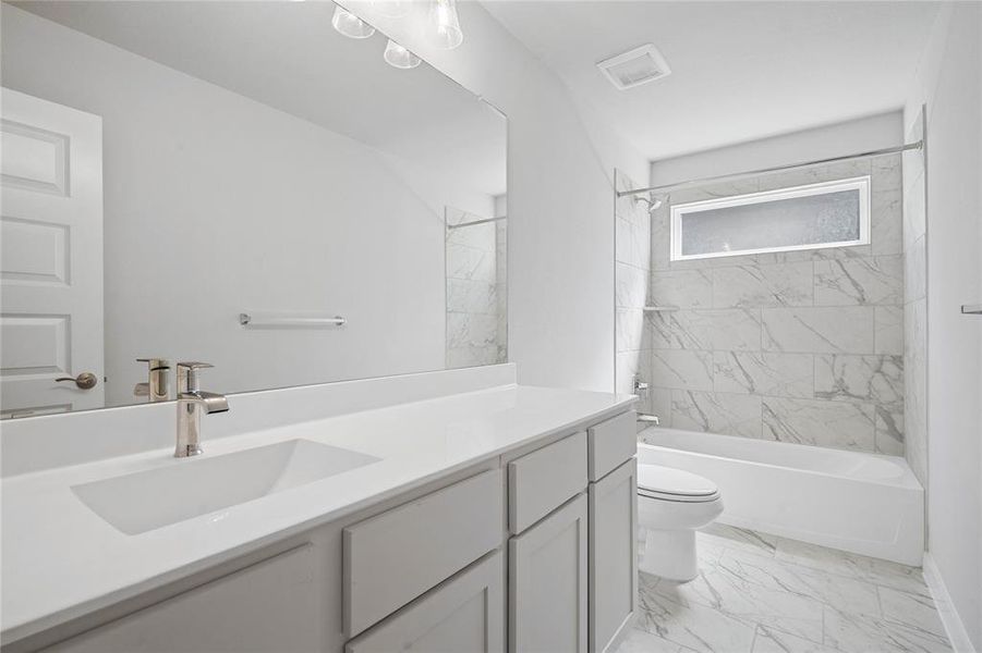 Secondary bath features tile flooring, bath/shower combo with tile surround, stained wood cabinets, beautiful light countertops, mirror, dark, sleek fixtures and modern finishes.