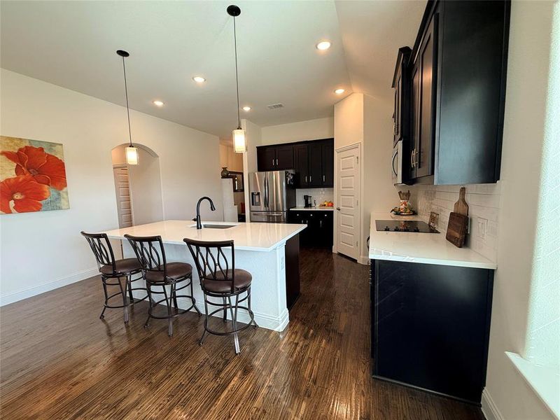 Kitchen featuring a large central island with white countertop, dark cabinetry, wood-finish flooring, recessed lighting, and pendant light fixtures Kitchen featuring a large central island with white countertop, dark cabinetry, wood-finish flooring, recessed lighting, and pendant light fixtures