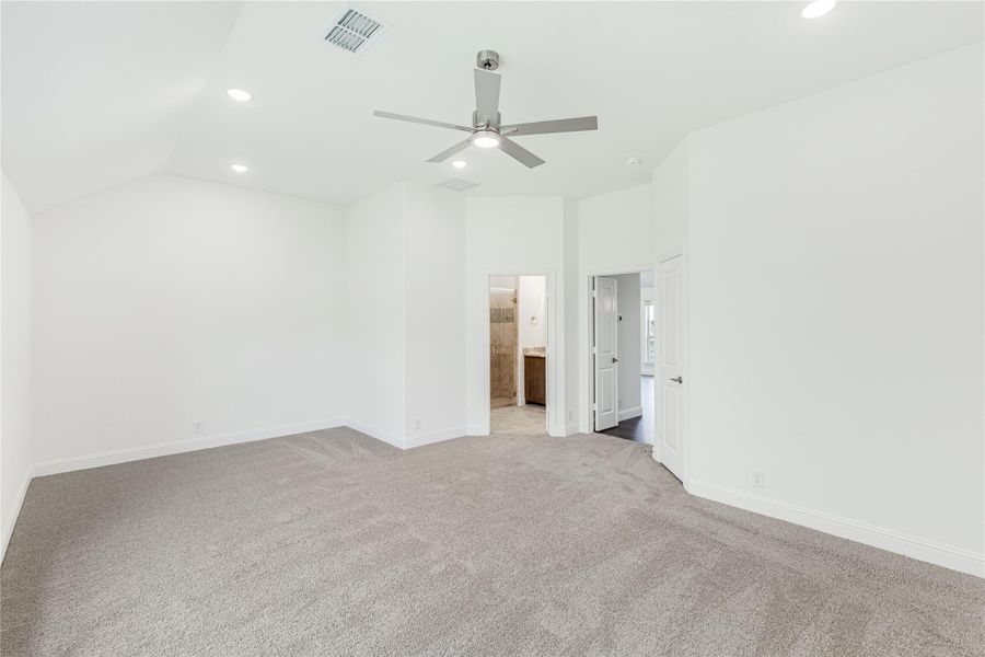 Empty master bedroom with vaulted ceiling, ceiling fan, carpet flooring, and ensuite bathroom doorway Empty master bedroom with vaulted ceiling, ceiling fan, carpet flooring, and ensuite bathroom doorway