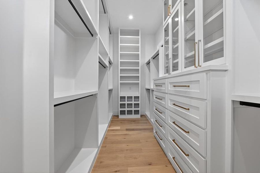 Custom closet system featuring white cabinetry, glass-front upper cabinets, built-in drawers with brass hardware, open shelving, and wood-finish flooring Custom closet system featuring white cabinetry, glass-front upper cabinets, built-in drawers with brass hardware, open shelving, and wood-finish flooring