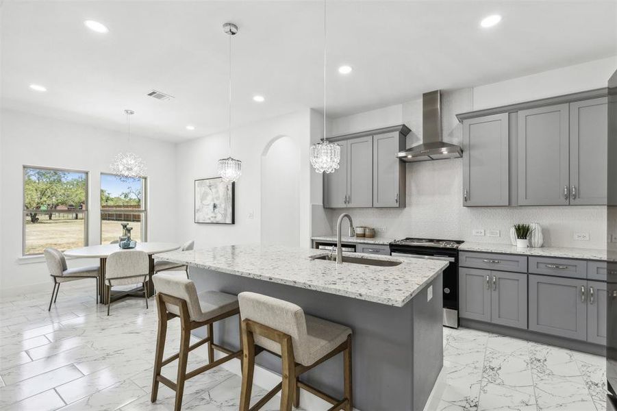 Kitchen with hanging light fixtures, an island with sink, light stone counters, wall chimney exhaust hood, and stainless steel range oven