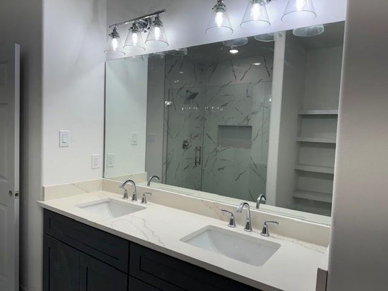 This is a modern bathroom featuring a double vanity with marble countertops, sleek dark cabinetry, and a large mirror with stylish overhead lighting. The shower area has glass doors and appears to have marble-style tiling.