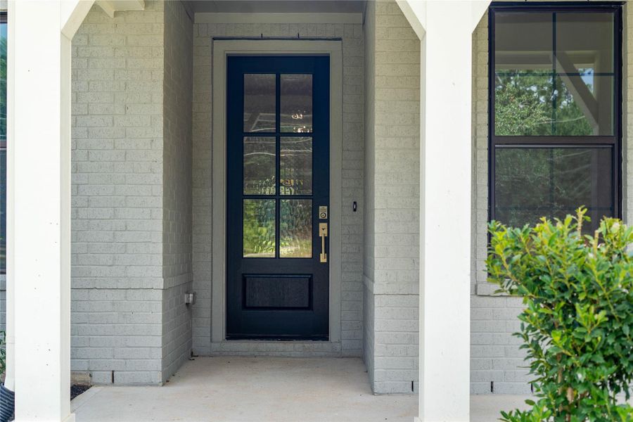 This charming front entryway boasts a premium 6-lite glass entry door painted in a timeless dark hue with elegant gold hardware. The light brick façade is accented by classic architectural pillars and a matching grid window design. Enjoy a seamless transition from the covered porch into this beautiful, light-filled property.