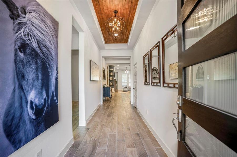 The floor is covered with elegant wood-look tile in natural tones, adding warmth and texture to the space. The ceiling features a recessed wooden accent panel with a geometric chandelier that provides modern lighting and a touch of sophistication.