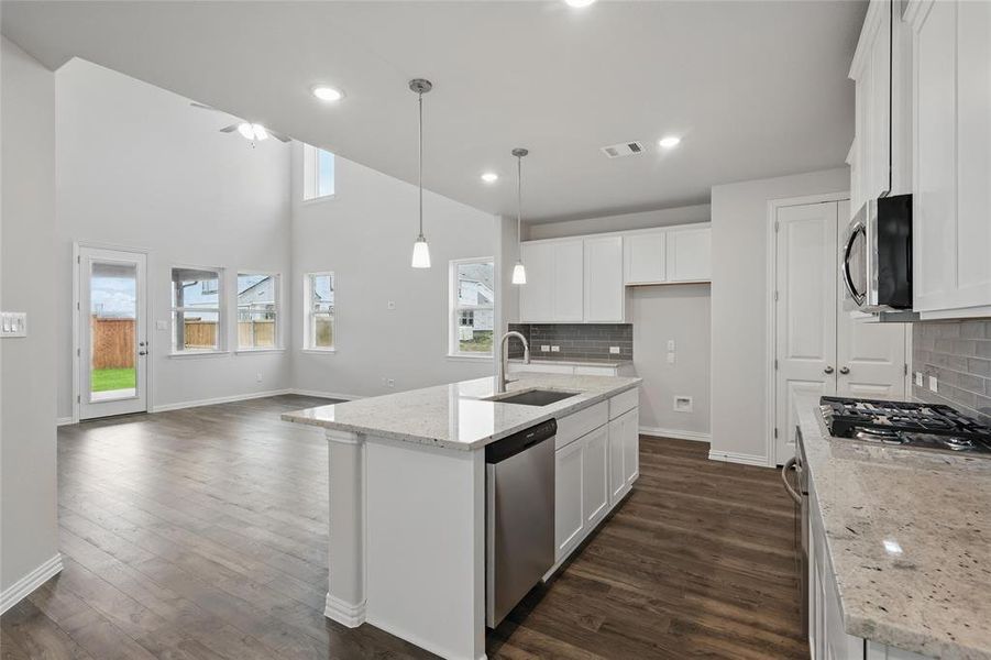 Kitchen featuring light stone counters, an island with sink, backsplash, decorative light fixtures, and dark wood-type flooring