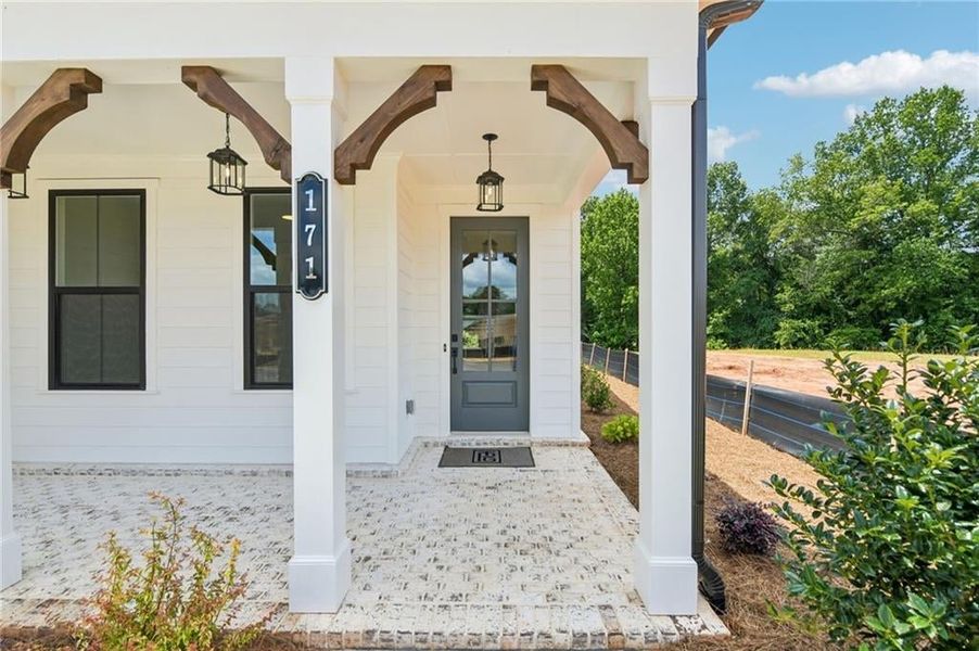 Welcoming front porch featuring whitewashed brick flooring, decorative wood corbels, and a dark gray entry door with glass inserts
